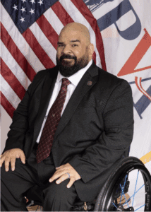 A man in a suit and tie sits in a wheelchair, posed in front of American flags and a banner. He has a beard, shaved head, and is smiling slightly, with his hands resting on his knees.